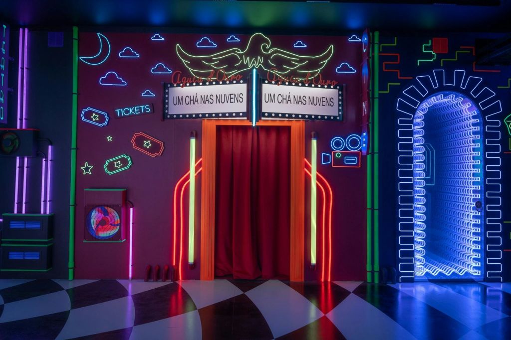 "Um Chá Nas Nuvens" 간판 옆에 빨간 커튼이 있는 네온 조명 입구는 다양한 네온 디자인으로 화려한 벽에 세워져 있습니다.