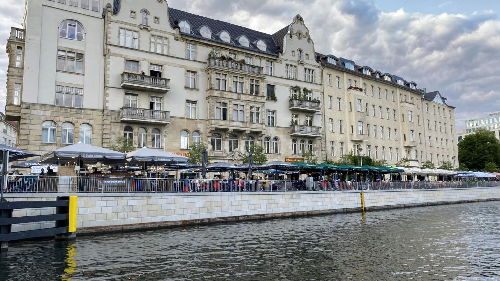 유서 깊은 유럽풍 건물이 우산 아래 야외 카페가 있는 강둑에 늘어서 있으며, 사람들이 식사를 하고 있습니다.