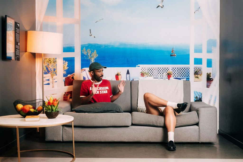 해안 테마의 배경이 있는 방의 회색 소파에 앉아 있는 두 사람, 근처에 꽃과 과일이 놓인 사이드 테이블.