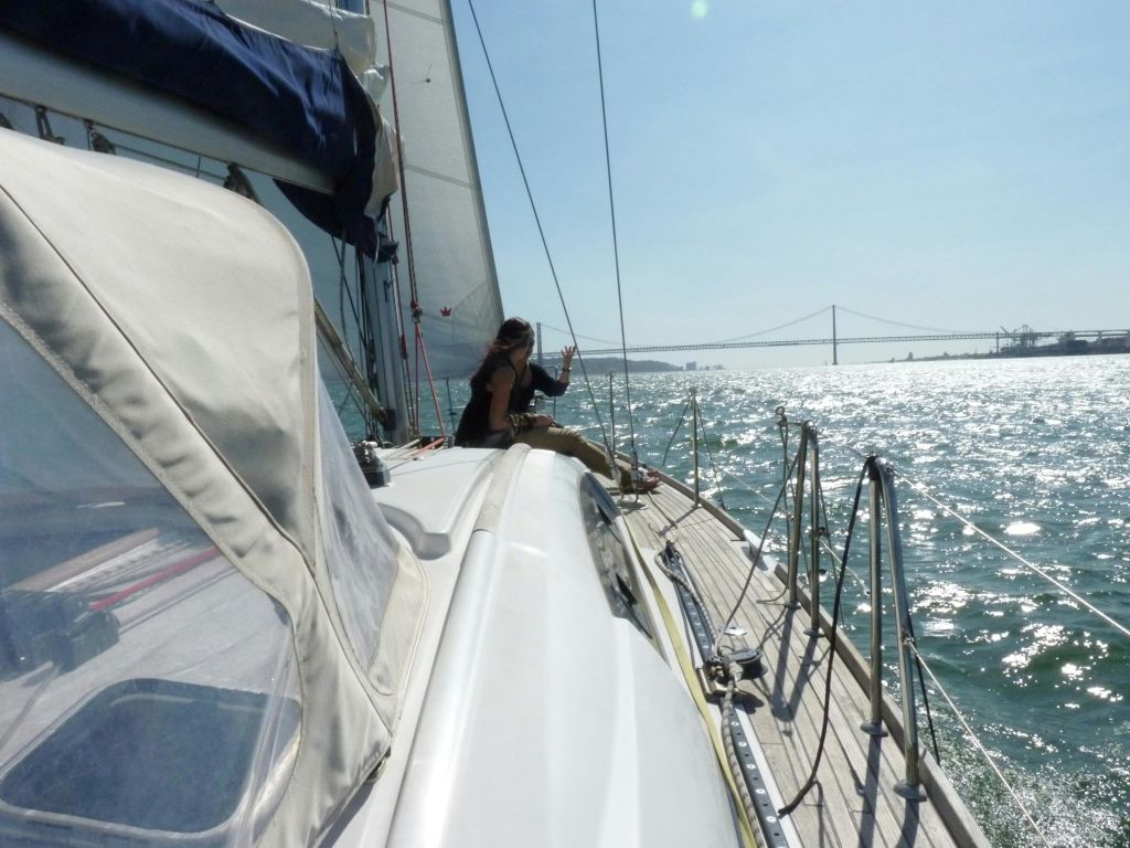 맑은 하늘 아래 바다와 먼 다리가 보이는 세일링 보트 갑판에 앉아 있는 사람.