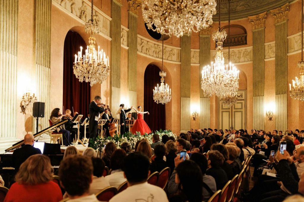 오케스트라가 무대에서 붉은 드레스를 입은 여성 댄서와 함께 관객에게 둘러싸인 샹들리에가 있는 그랜드 홀에서 공연합니다.