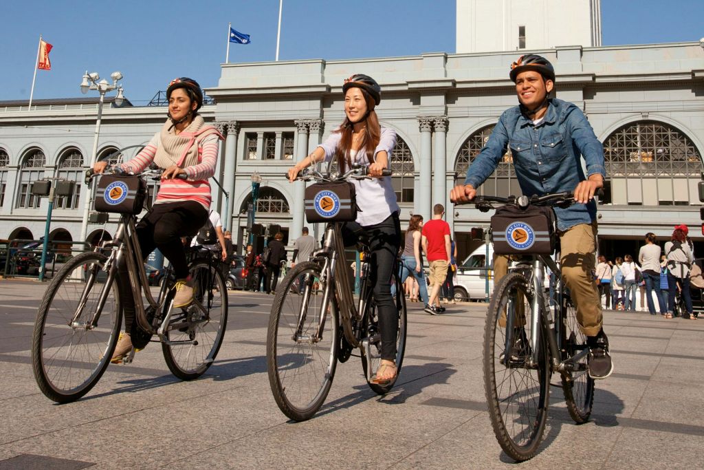 아치형 창문과 시계탑이 있는 커다란 건물 앞에서 자전거를 타고 있는 헬멧을 쓴 두 사람.