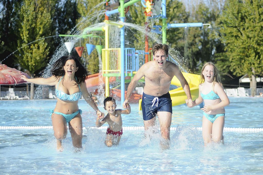4인 가족이 수영복을 입고 미끄럼틀과 분수가 있는 야외 워터파크에서 얕은 물 속을 거닐고 있습니다.