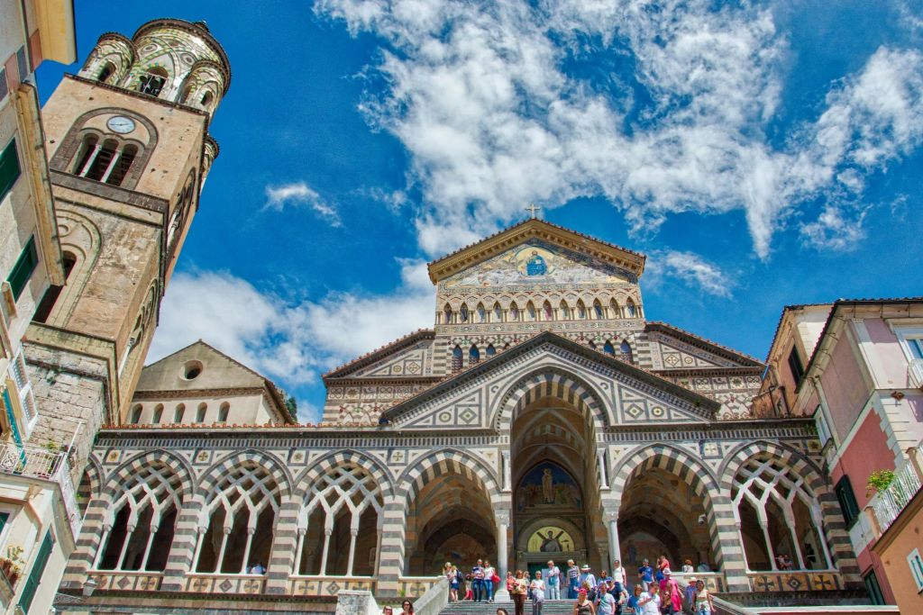 정교한 줄무늬 아치와 푸른 하늘 아래 장식된 정면이 있는 대성당 앞에 사람들이 모여 있습니다.