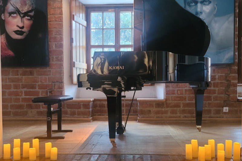검은색 카와이 피아노가 놓인 방, 벤치, 초상화가 놓인 벽돌 벽, 바닥에 놓인 촛불, 나무 셔터가 달린 창문.