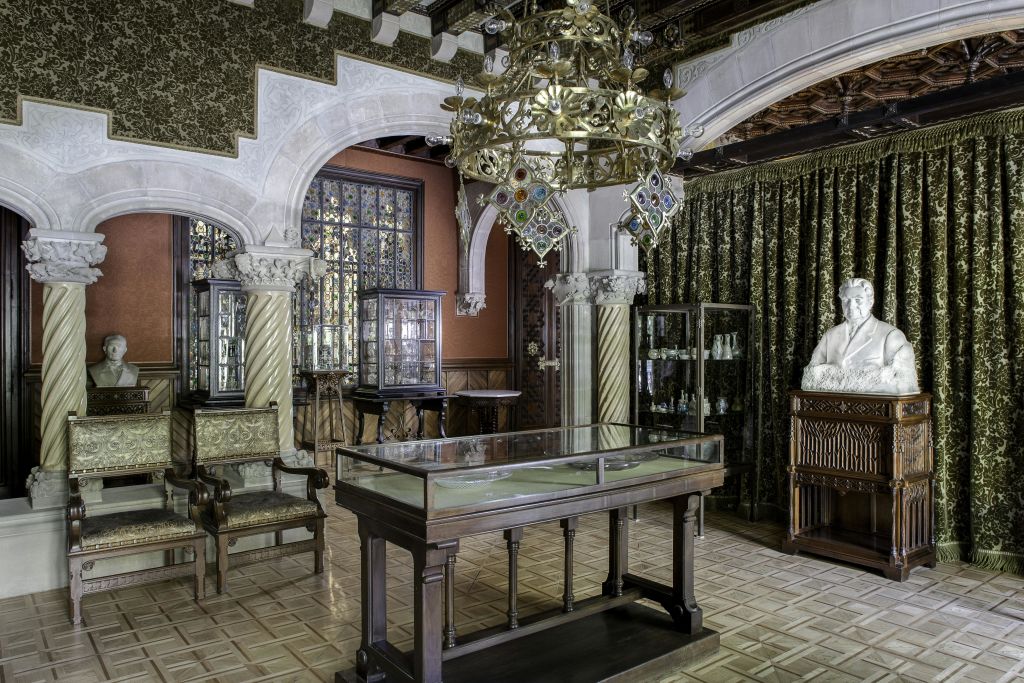 화려한 샹들리에, 유리 물품이 놓인 진열장, 조각된 의자, 대리석 기둥, 나무 받침대에 새겨진 하얀 흉상이 있는 호화로운 객실입니다.