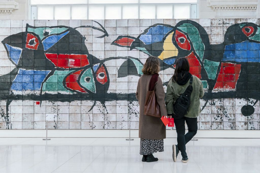 밝은 실내 공간에서 타일로 장식된 다채로운 추상 벽화 앞에 두 사람이 서 있습니다.