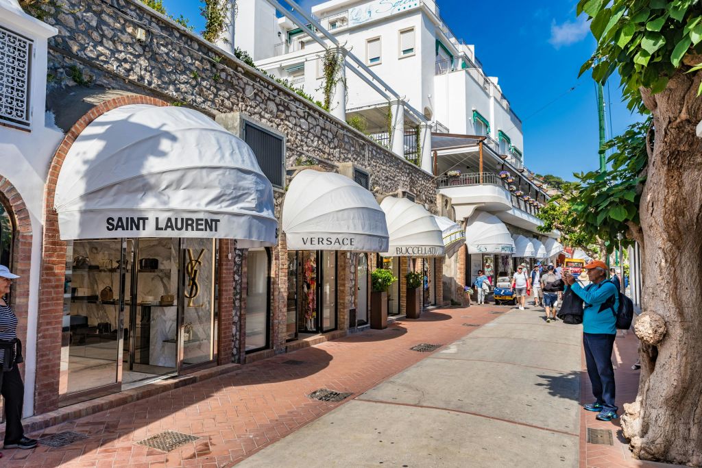 생 로랑, 베르사체 및 기타 부티크가 있는 야외 쇼핑 지역. 쇼핑객들은 맑은 하늘 아래 포장된 통로를 따라 걷습니다.