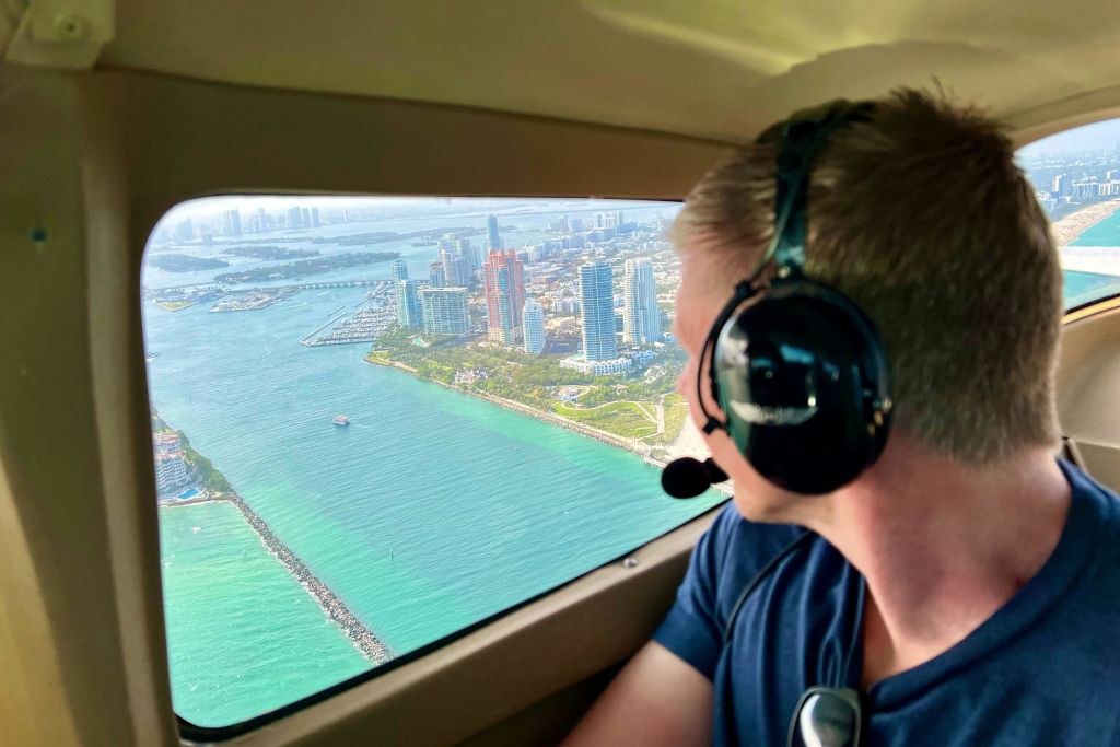 헤드셋을 착용한 사람이 청록색 바다를 따라 높은 건물과 해안선이 있는 도시의 작은 비행기 창문을 내다보고 있습니다.