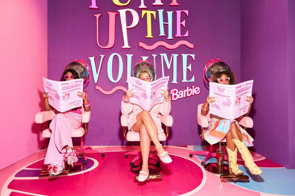 헤어드라이어 아래 미용실 의자에 앉아 분홍색 신문을 읽는 세 명의 여성. 뒤쪽 벽에 화려한 글씨로 "PUMP UP THE VOLUME".