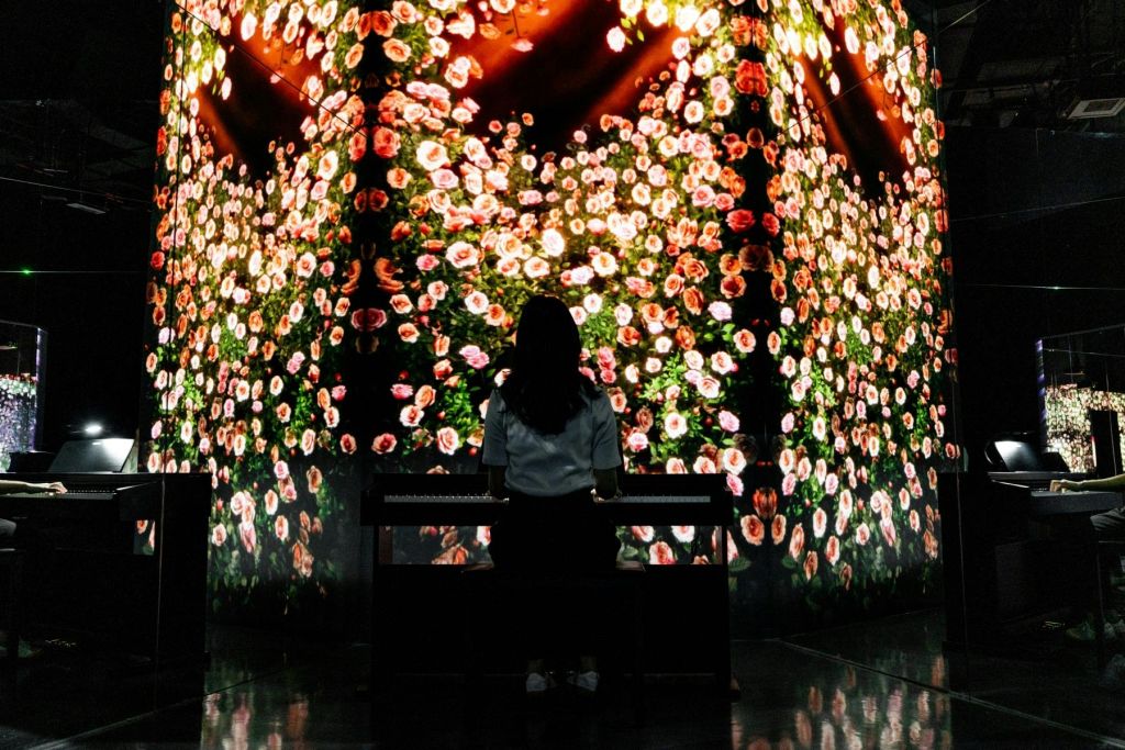 한 사람이 분홍색과 흰색 꽃과 따뜻한 조명으로 활기찬 꽃무늬를 보여주는 대형 스크린을 마주한 피아노에 앉아 있습니다.