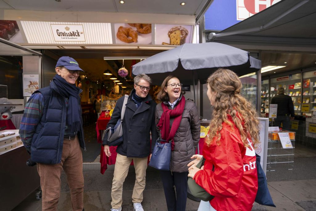 우산과 간판이 배경으로 보이는 베이커리 밖에서 성인 4명이 상호작용하며 미소를 짓고 있습니다.