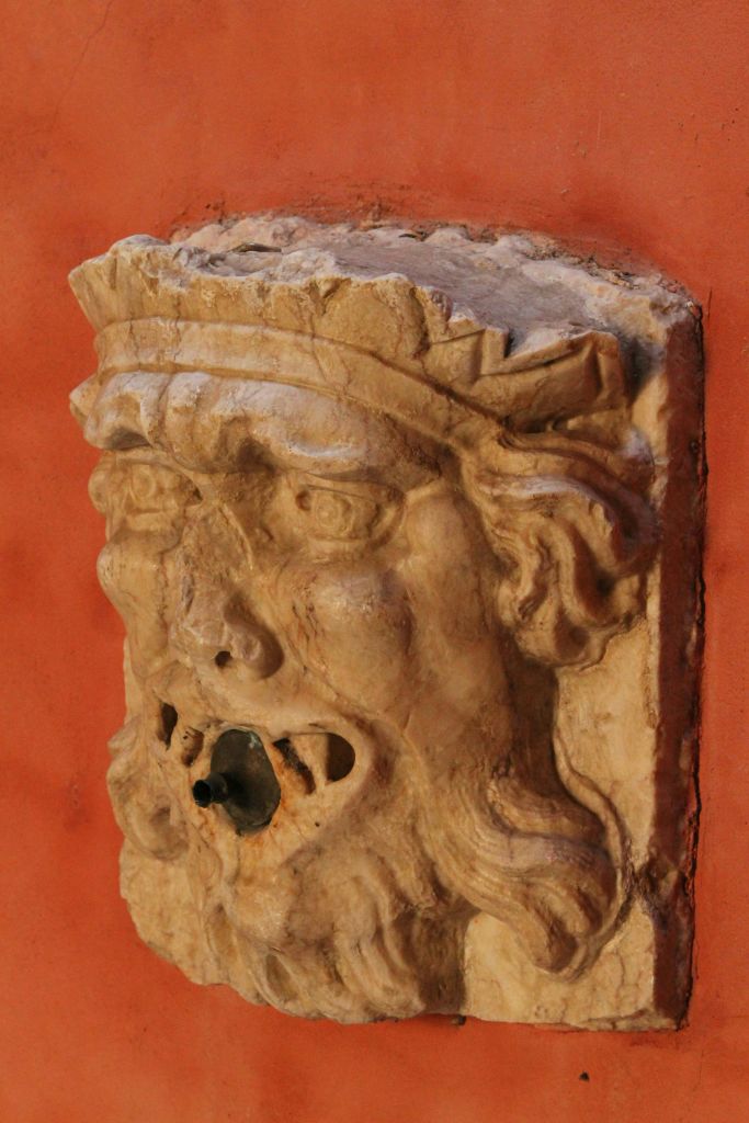 수염이 무성한 얼굴, 마모된 디테일, 입을 벌린 풍화된 석조 분수대가 붉게 물든 벽에 세워져 있습니다.