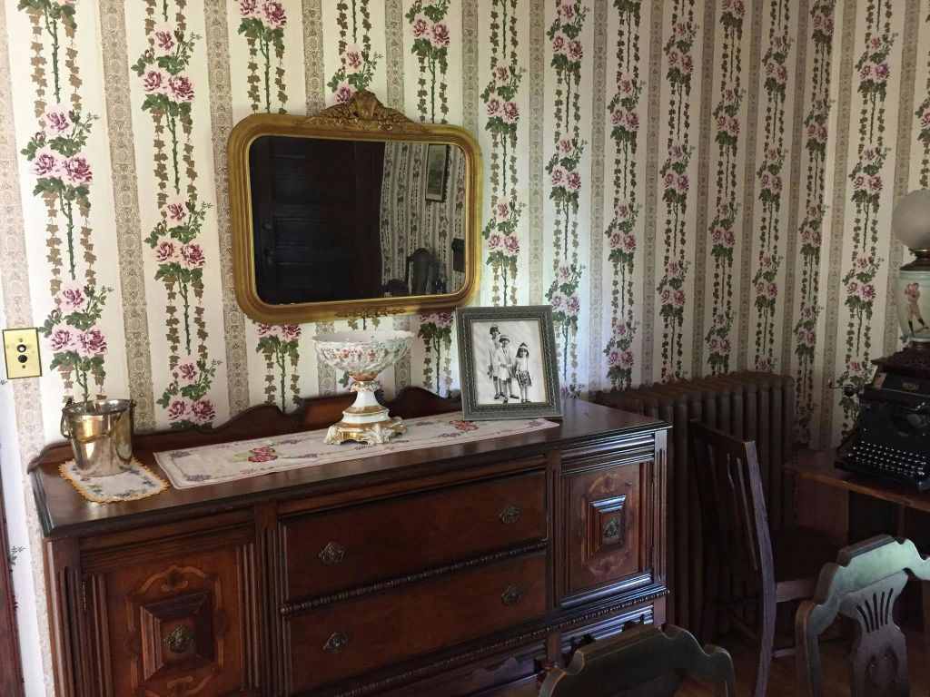 꽃무늬 벽지가 있는 방, 도일리가 있는 나무 서랍장, 흑백 액자 사진, 장식용 그릇, 커다란 거울이 있는 방.