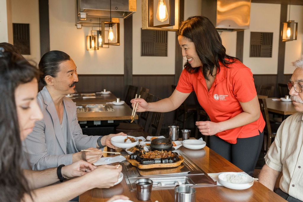 저명한 콧수염을 가진 젊은 남성에게 한국식 바비큐를 제공하는 밝은 빨간색 셔츠를 입은 밴쿠버 미식가 투어 가이드.