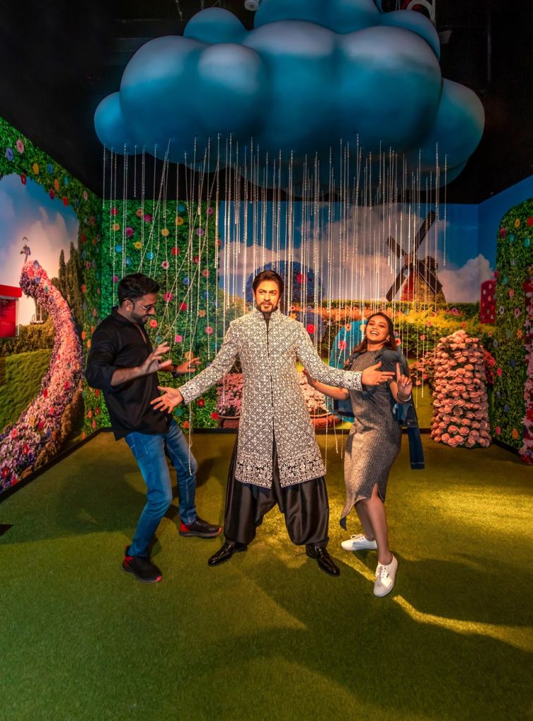 활기찬 정원과 시골 풍경으로 꾸며진 방에서 구름 아래에서 세 사람이 춤을 추며 포즈를 취하고 있습니다.