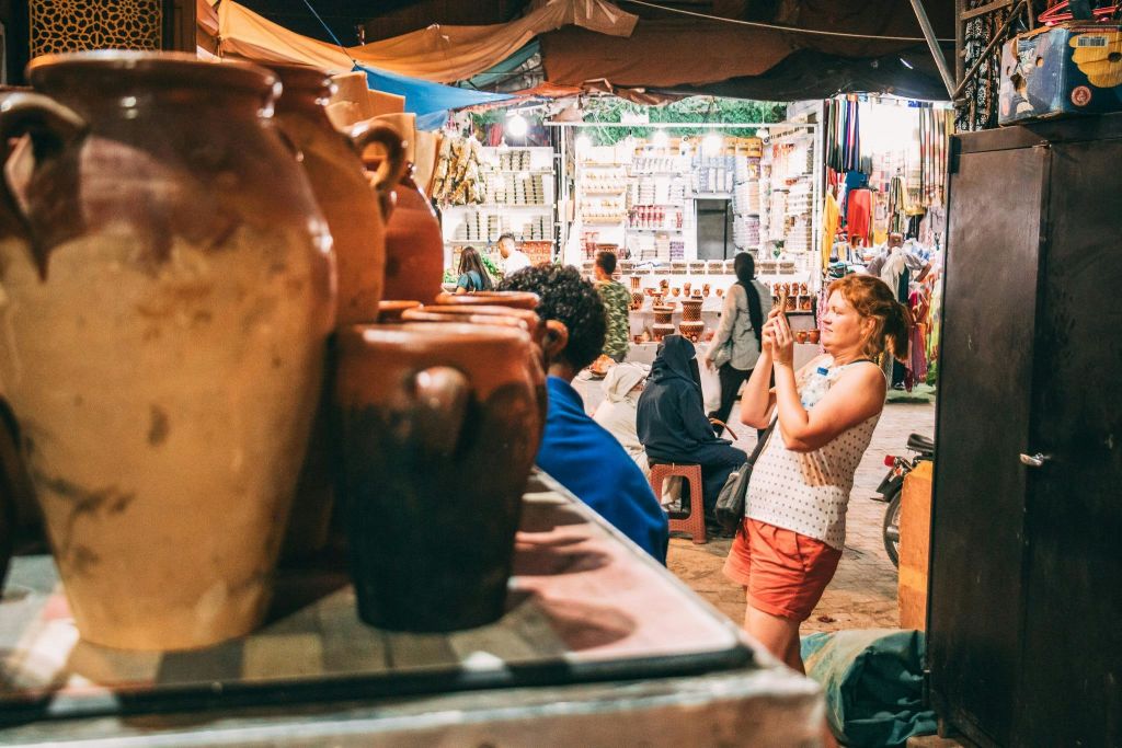 여름 옷을 입은 한 여성이 도자기, 직물, 다양한 상품으로 가득한 시끌벅적한 시장에서 사진을 찍고 있습니다.