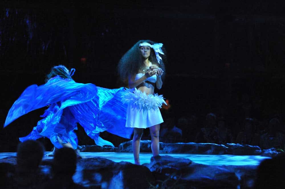 흰색 의상과 머리띠를 한 여성이 무대 위에 서서 물건을 들고 있고, 파란색 옷을 입은 다른 사람이 뒤에서 움직입니다.