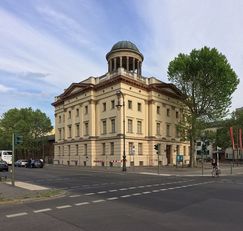 나무와 자전거 타는 사람, 주차된 차량으로 둘러싸인 교차로에 돔형 옥상이 있는 신고전주의 양식의 건물입니다.
