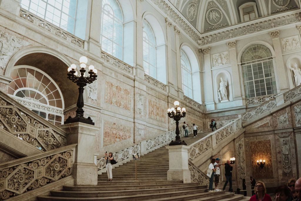 높은 창문, 장식용 램프, 조각상, 걷거나 서 있는 여러 사람이 있는 화려한 건물의 웅장한 계단입니다.