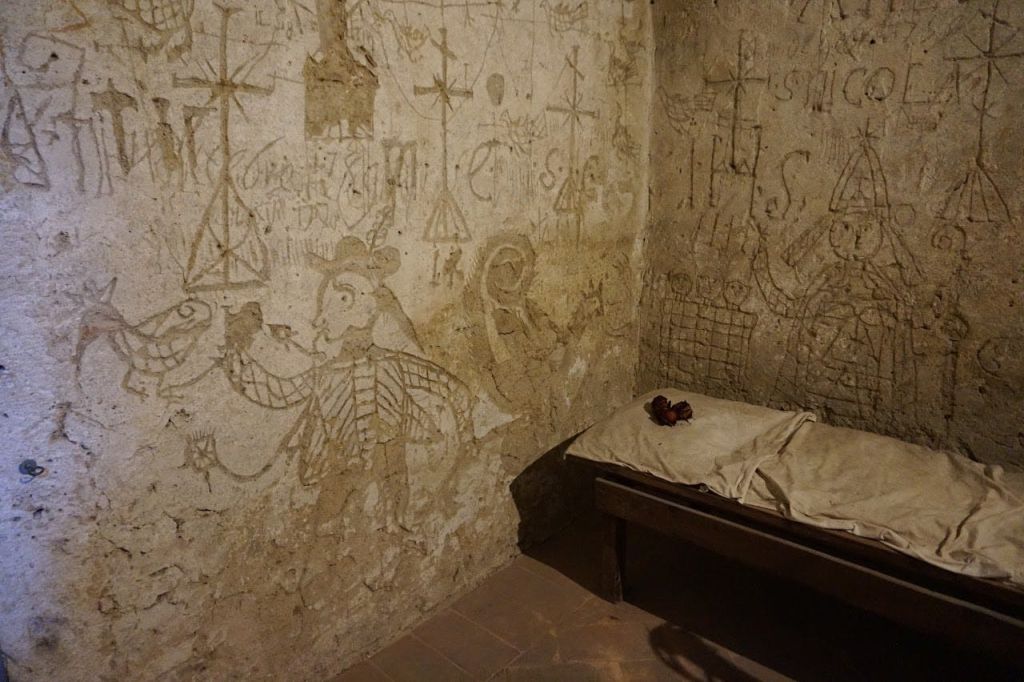 오래된 도면과 조각으로 덮인 벽이 있는 작은 방입니다. 마모된 침구와 빨간색 천 다발이 놓인 심플한 침대가 있습니다.