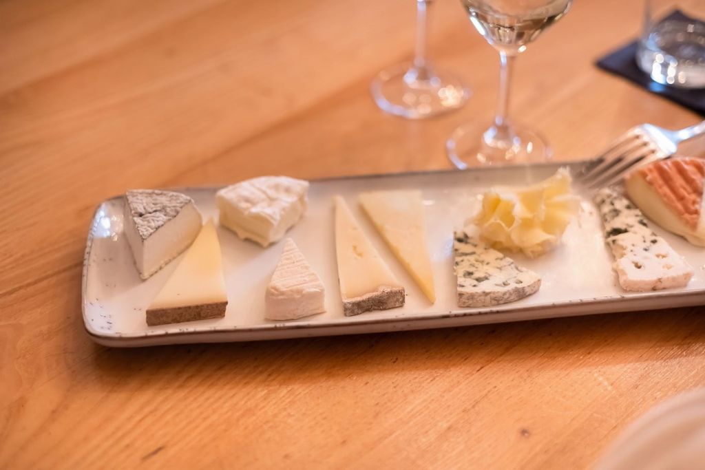 나무 테이블 위에 모듬 치즈 슬라이스가 놓인 직사각형 접시, 두 개의 와인 잔이 배경으로 놓여 있습니다.