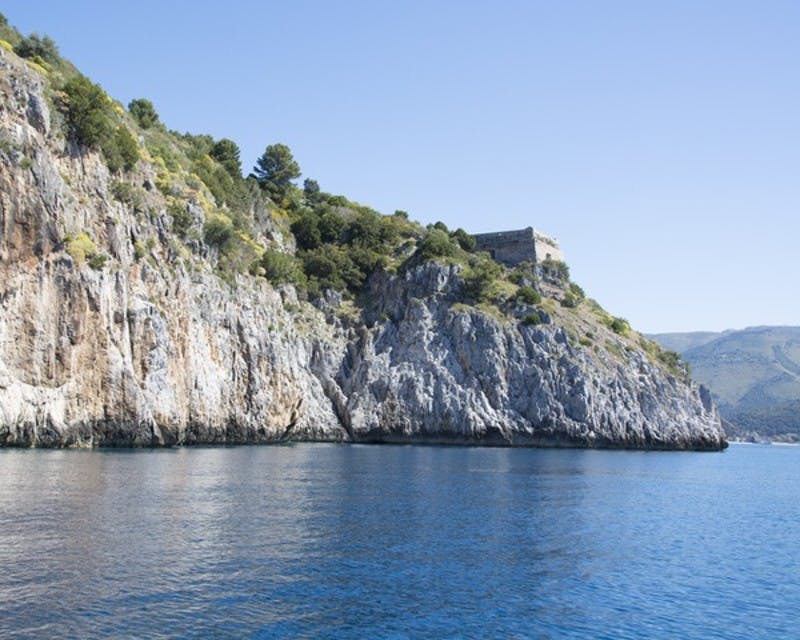 푸른 초목이 우거진 바위 해안 절벽과 청명한 하늘 아래 잔잔한 푸른 물 옆에 있는 꼭대기에는 작은 석조 건물이 있습니다.