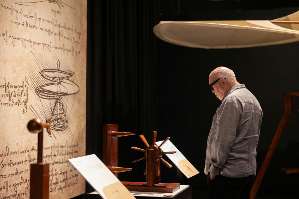 회색 셔츠를 입은 한 남성이 희미하게 빛나는 전시물에 전시된 나무 모형을 바라보고 있습니다. 천장에 커다란 구조물이 걸려 있습니다.