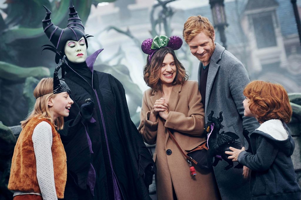 가족이 테마 공원에서 말레피센트 캐릭터와 함께 포즈를 취하며 웃고 있습니다. 소녀와 소년은 좋아하는 캐릭터와 어울리는 머리띠를 착용합니다.