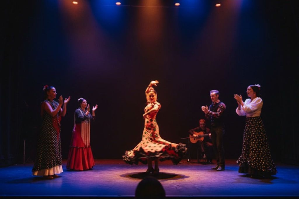 빨간색과 검은색 폴카 도트 드레스를 입은 플라멩코 댄서가 무대 조명 아래에서 연주자와 클래퍼를 배경으로 공연을 펼칩니다.