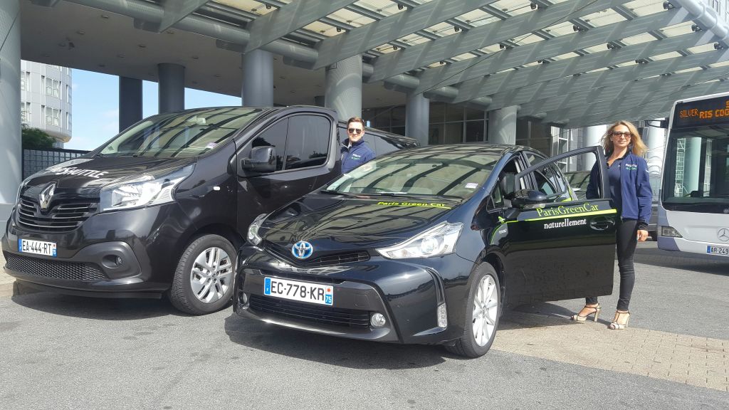 재킷을 입은 두 명의 여성이 "파리 그린카" 표지판이 있는 검은색 자동차 옆에 서 있습니다. 버스가 배경에 있습니다.