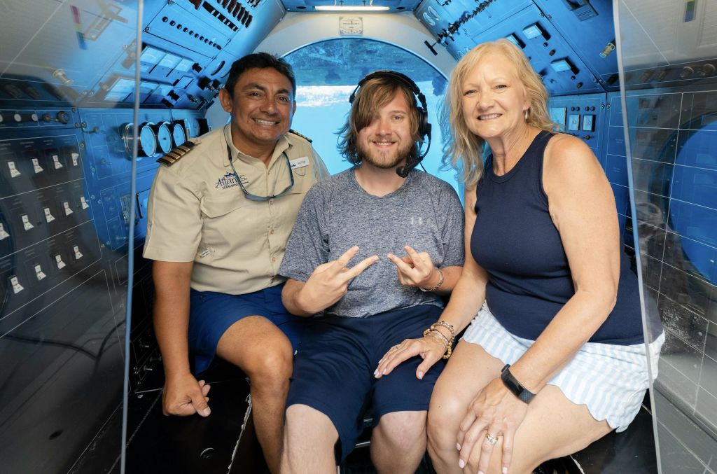 파란색 잠수함 안에 세 사람이 앉아 있습니다. 한 사람은 헤드폰을 착용하고, 다른 한 사람은 미소를 짓고, 한 여성은 민소매 상의를 입고 있습니다.