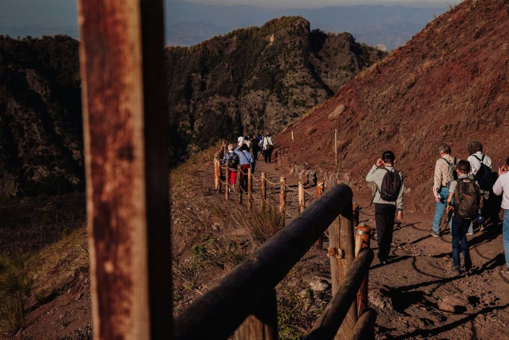 등산객들은 맑은 하늘 아래 언덕과 나무를 배경으로 나무 난간으로 둘러싸인 흙길을 따라 걷습니다.