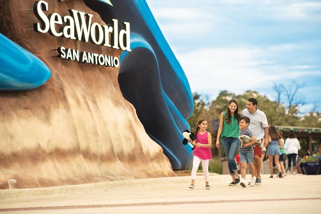 화창한 날 대형 "SeaWorld San Antonio" 표지판 근처에서 4인 가족이 함께 산책하며 다른 방문객들을 배경으로 이야기를 나눕니다.