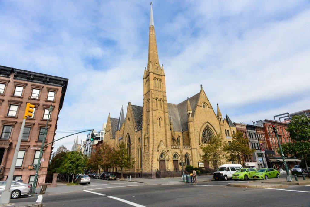 주차된 자동차와 보행자로 둘러싸인 도시 거리 모퉁이에 높은 첨탑이 있는 대형 석조 교회.