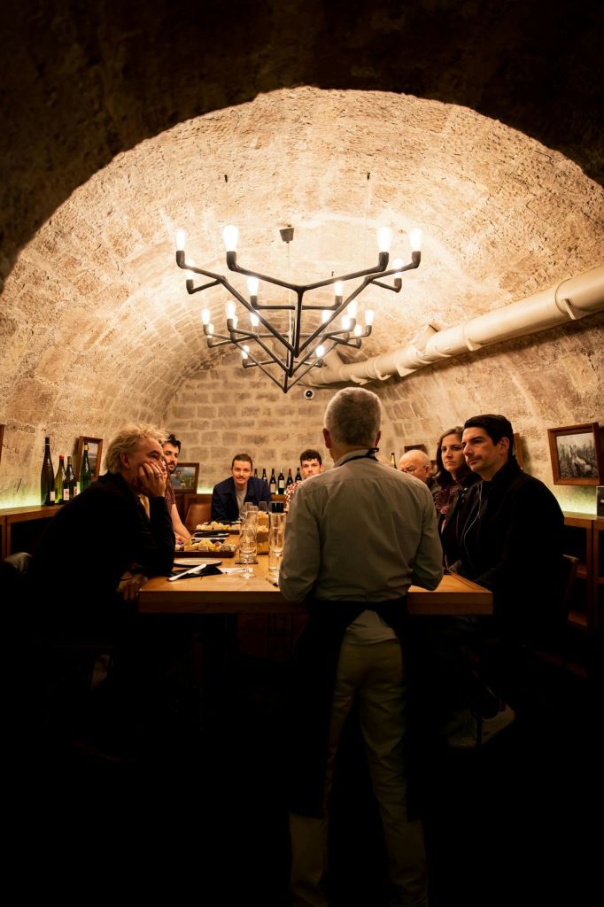 돌로 장식된 방의 식탁에 한 무리의 사람들이 앉아 있고, 웨이터가 테이블에 서 있습니다. 위에 샹들리에가 있습니다.