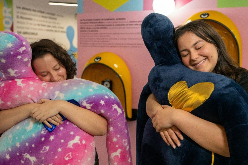 화려한 벽과 노란색 설비가 있는 방에서 한 사람이 금색 하트가 있는 커다란 파란색 봉제 장난감을 끌어안고 눈을 감은 채 미소를 짓고 있습니다.