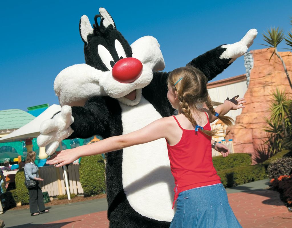 맑고 푸른 하늘 아래에서 어린 소녀가 고양이 실베스터 의상을 입은 사람을 향해 팔을 뻗고 있습니다.