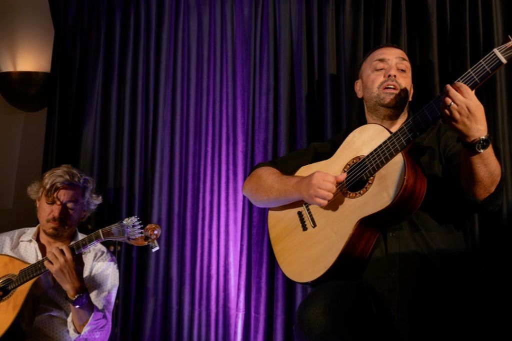 어두운 커튼과 보라색 조명이 있는 무대에서 두 남자가 어쿠스틱 기타를 연주하고 있습니다.