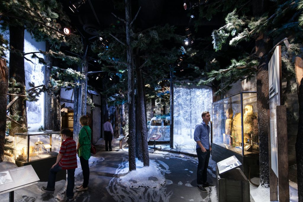 박물관에 있는 한 사람이 눈으로 덮인 나무를 전시하고 있으며, 겨울 의류와 유물이 담긴 유리 진열 케이스를 보고 있습니다.