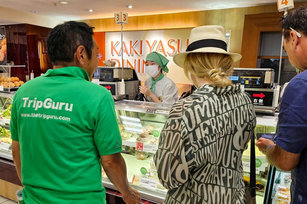 두 사람이 푸드 카운터에 서 있습니다. 한 명은 초록색 "TripGuru" 셔츠를 입고, 다른 한 명은 모자를 쓰고 있습니다. 유니폼을 입은 직원이 보조합니다. 백그라운드에서 "Kakiyasu Dining" 사인.