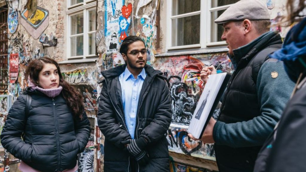 한 사람이 사진을 들고 있는 그래피티로 덮인 벽 앞에 세 사람이 서 있습니다.