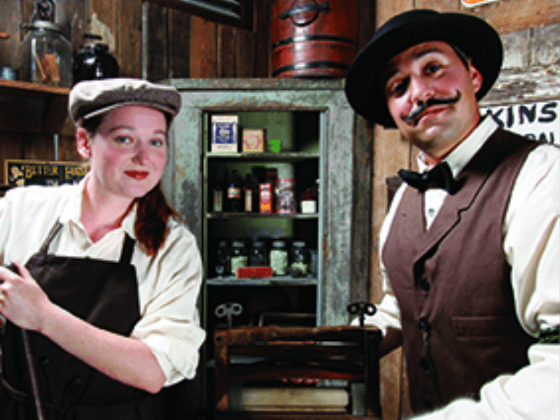 빈티지 복장을 한 두 사람이 다양한 항아리와 상자가 놓인 나무 선반 앞에 서 있습니다.