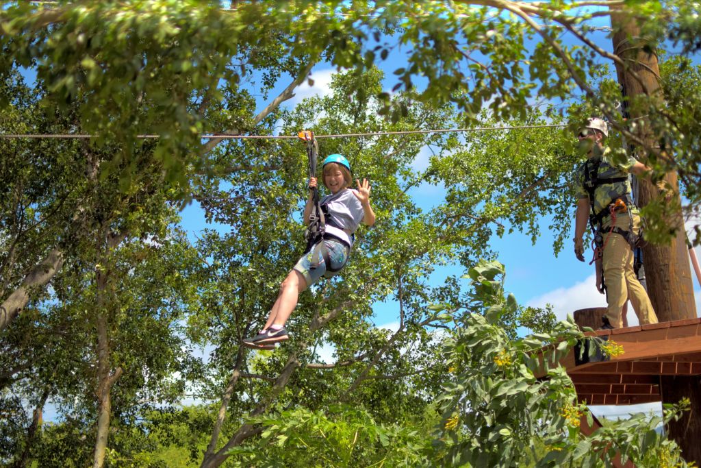 헬멧과 하네스를 착용한 사람이 숲을 가로질러 집라인 케이블을 잡고 미소를 짓고 있습니다.
