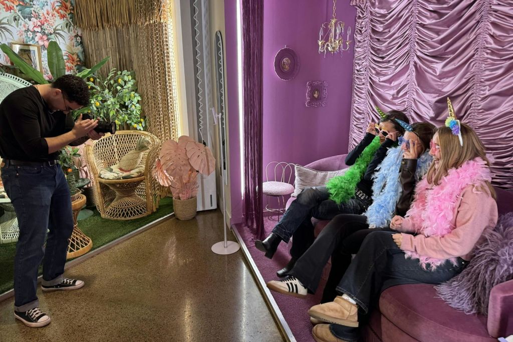 화려한 스카프와 유니콘 머리띠를 두른 세 사람이 선명한 보라색 인테리어가 돋보이는 방의 보라색 소파에 앉아 있습니다.