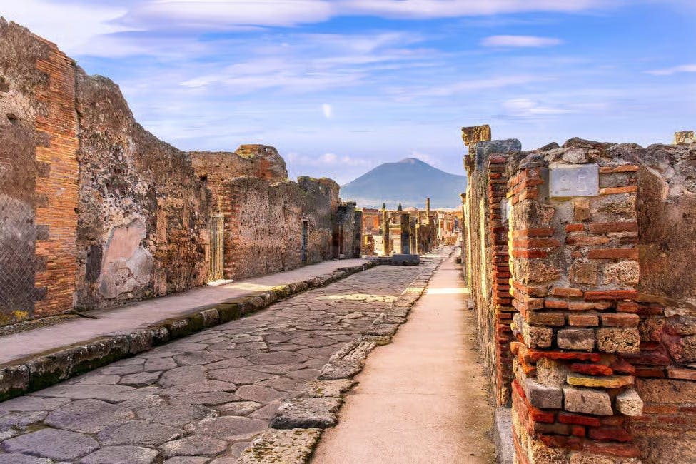 푸른 하늘 아래 돌로 포장된 거리와 침식된 벽이 있는 고대 유적지로, 저 멀리 산이 배경으로 보입니다.