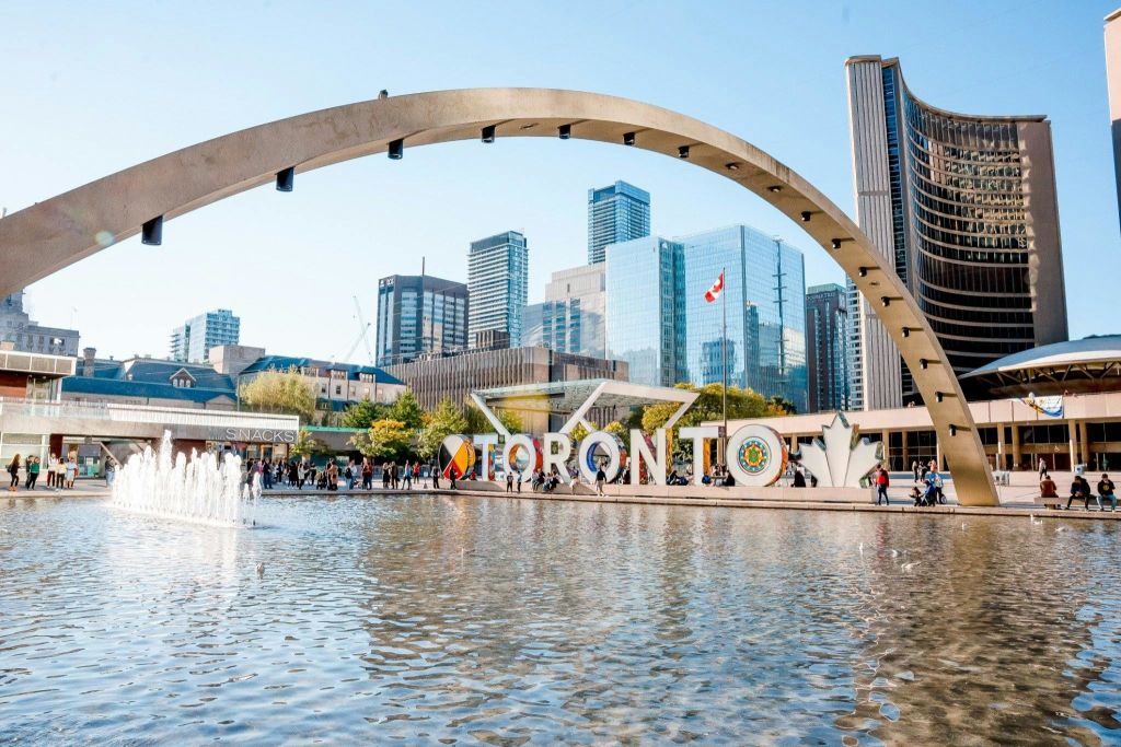 토론토의 네이선 필립스 스퀘어 (Nathan Phillips Square) 에는 분수, "토론토" 표지판, 맑은 하늘 아래 현대적인 건물이 있습니다.