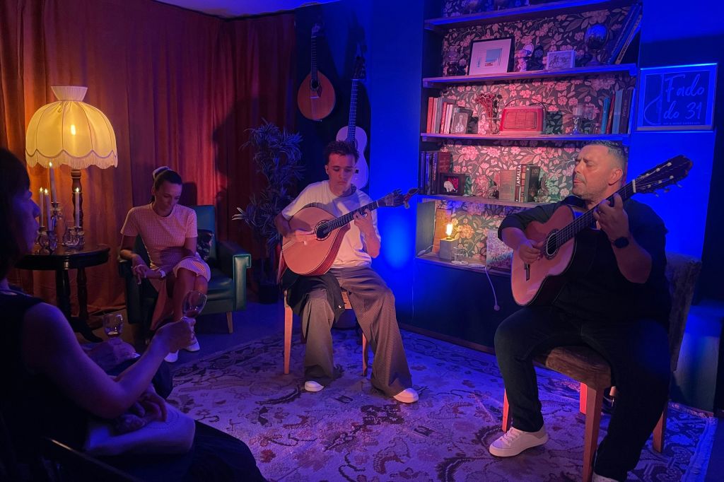 파란색과 보라색 조명 아래 선반, 책, 장식이 있는 아늑한 방에서 어쿠스틱 기타를 연주하는 두 사람.