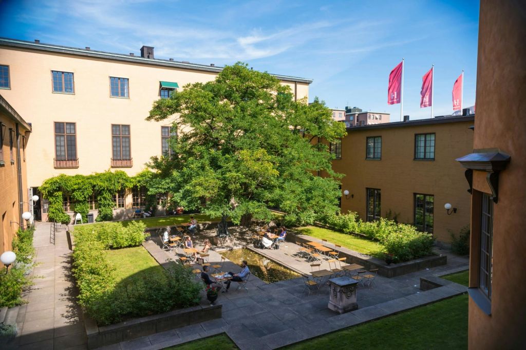 붉은 깃발이 보이는 푸른 하늘 아래 노란색 건물로 둘러싸인 나무가 있는 마당, 테이블에 앉아 있는 사람들.