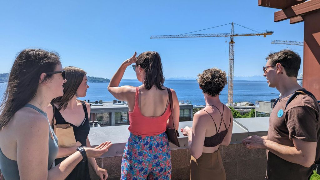 건축 크레인과 물을 배경으로 도시 풍경이 내려다보이는 발코니에 세 사람이 서 있습니다.